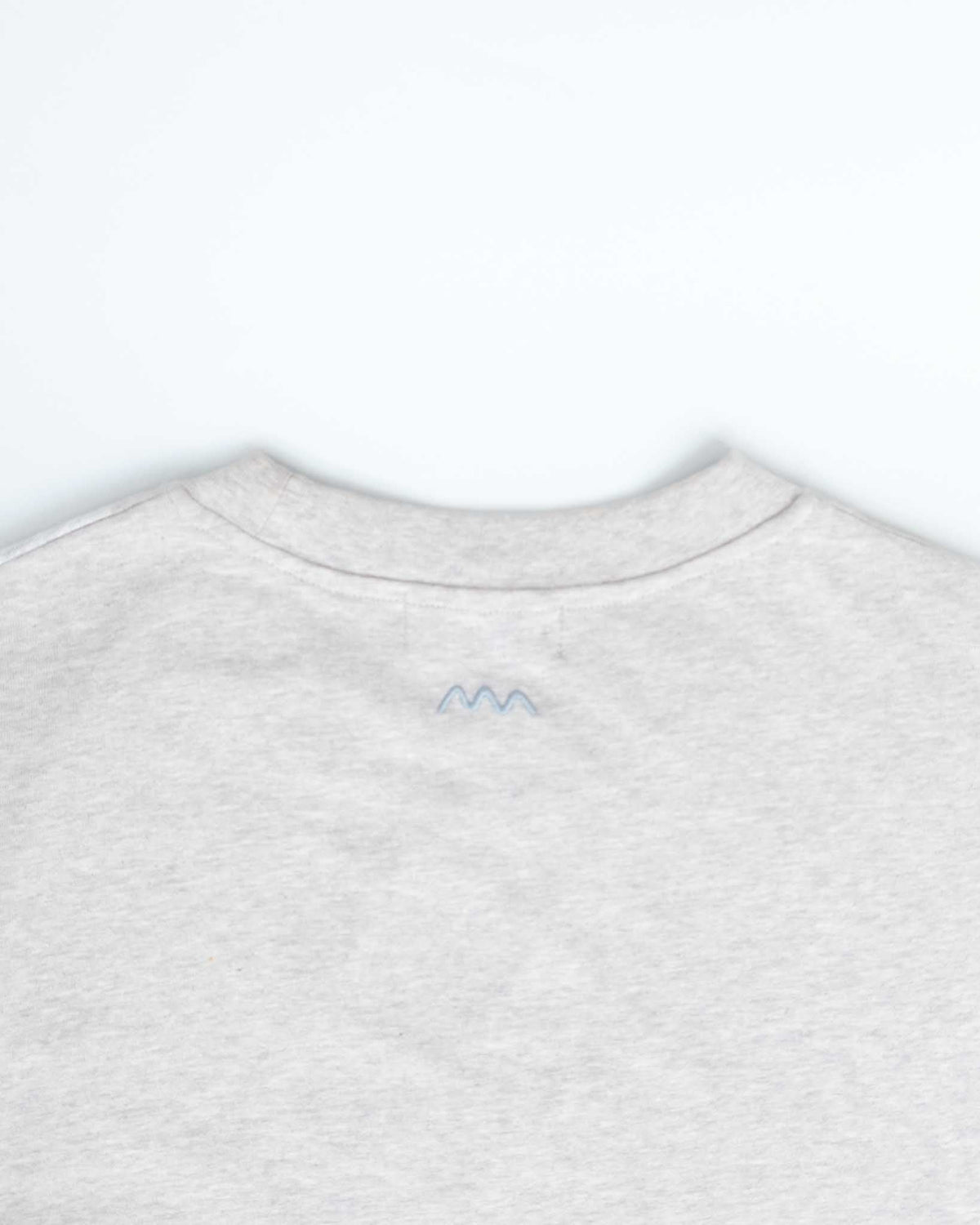 Close-up of the back of a light heather gray crewneck sweatshirt, featuring a small blue embroidered mountain-like logo centered below the collar.