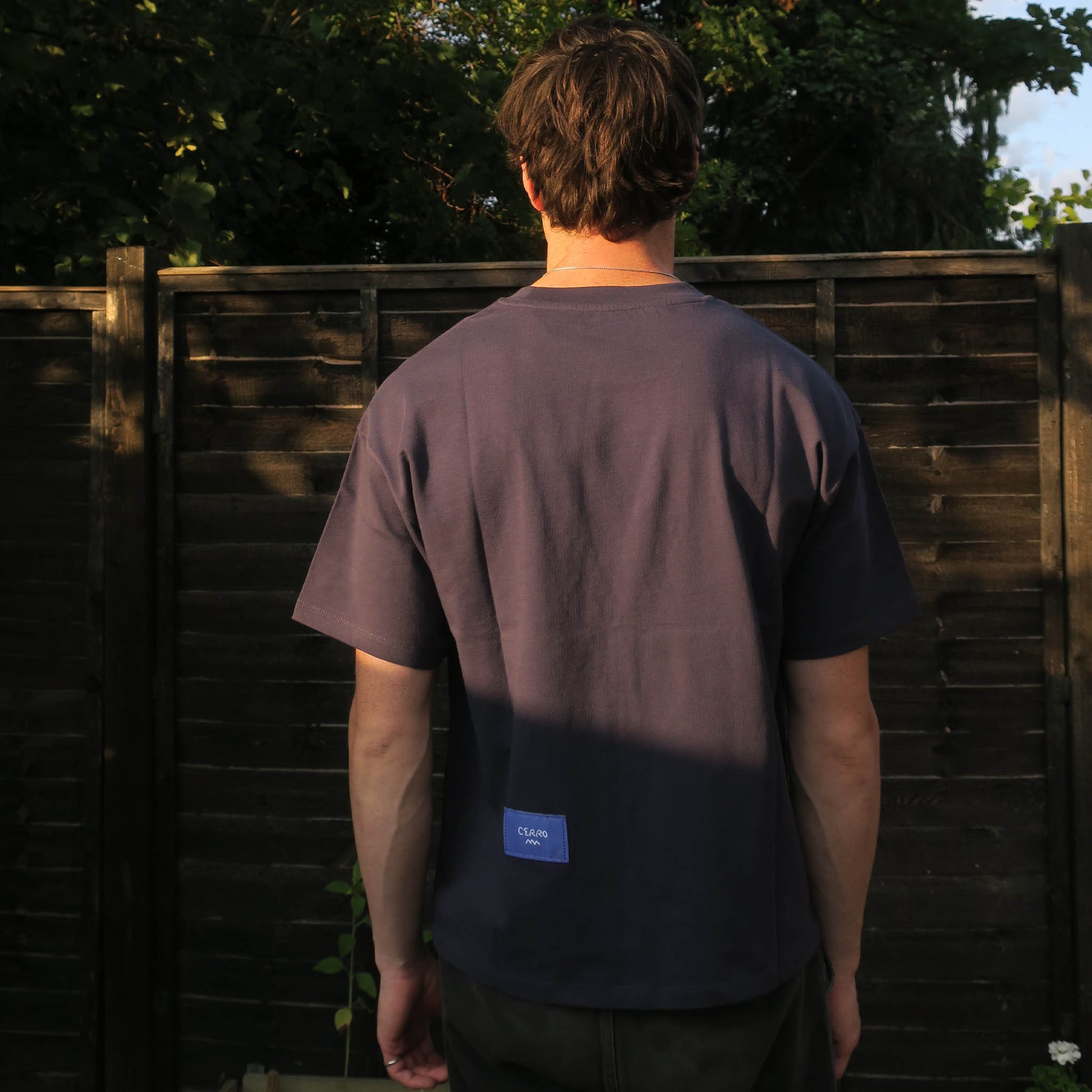 Person wearing a dark t-shirt with a logo on the back, standing in front of a wooden fence.