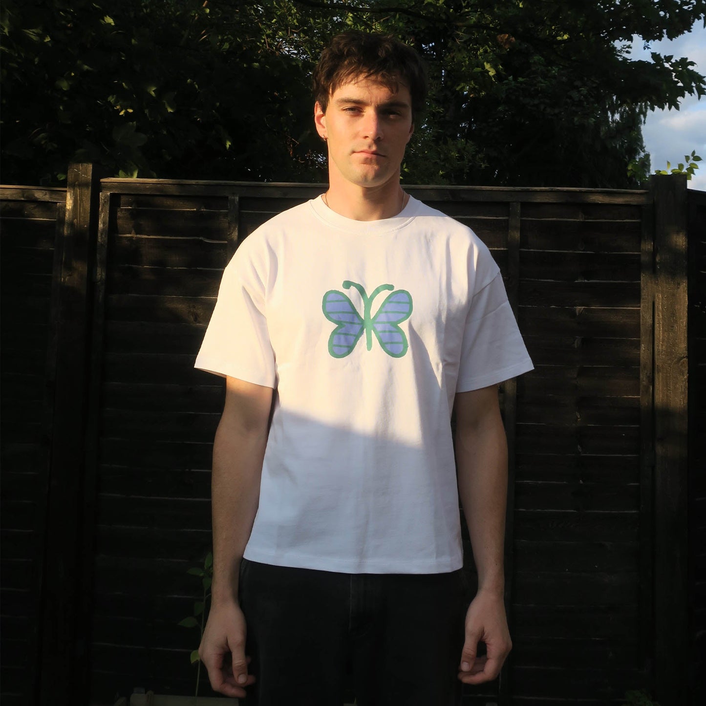 Person wearing a white t-shirt with a butterfly design in front of a wooden fence.