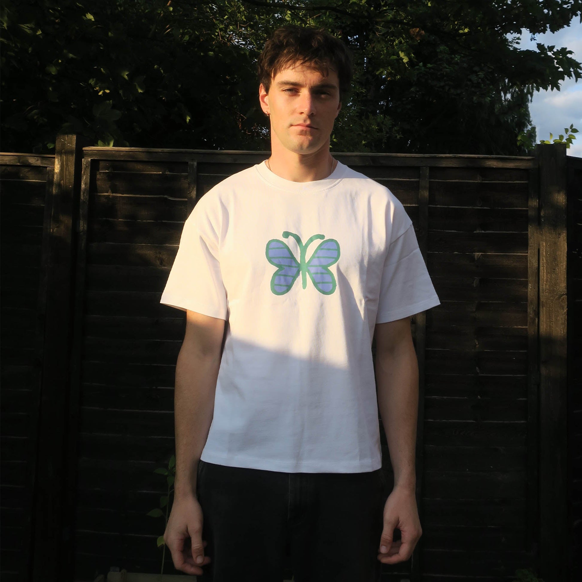 Person wearing a white t-shirt with a butterfly design in front of a wooden fence.