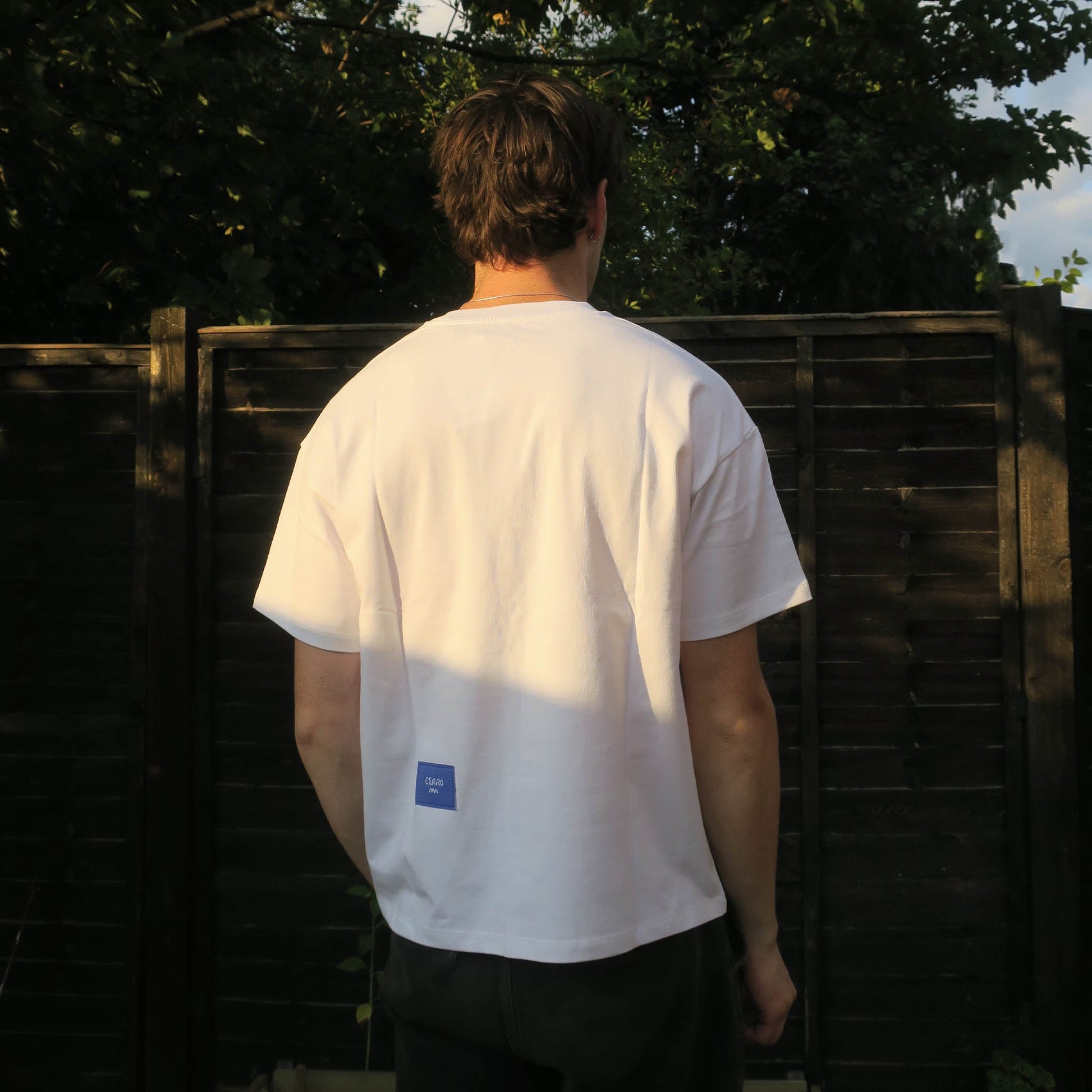 Person wearing a white t-shirt with a small blue logo, standing in front of a wooden fence.