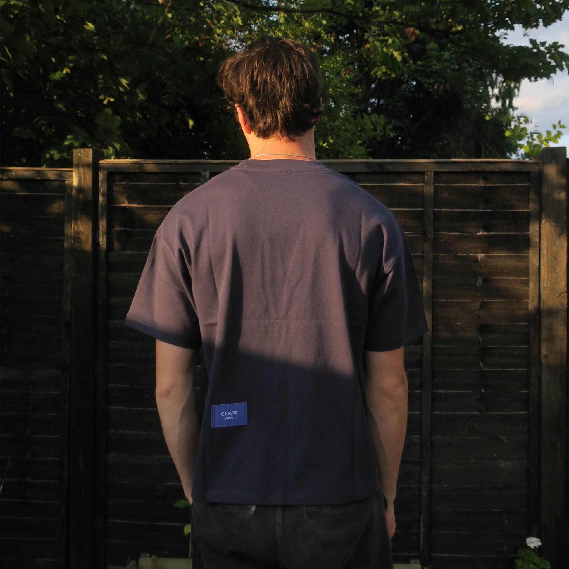 Person wearing a dark blue t-shirt with a blue logo on the, standing in front of a wooden fence.
