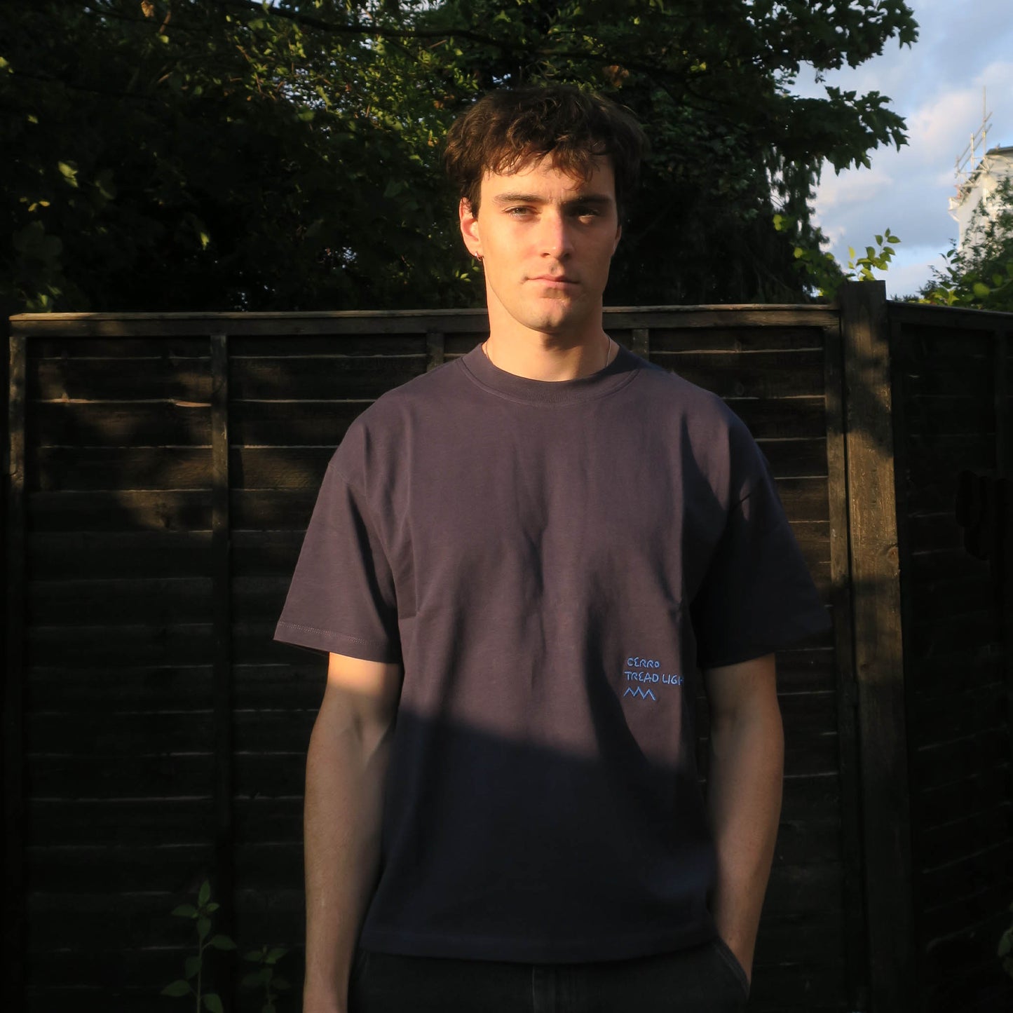 Person wearing a navy t-shirt with embroidered blue text, standing in front of a wooden fence with trees in the background.