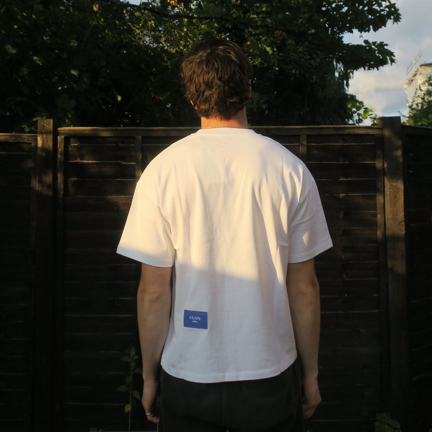 Person wearing a white t-shirt with a blue logo standing in front of a wooden fence.