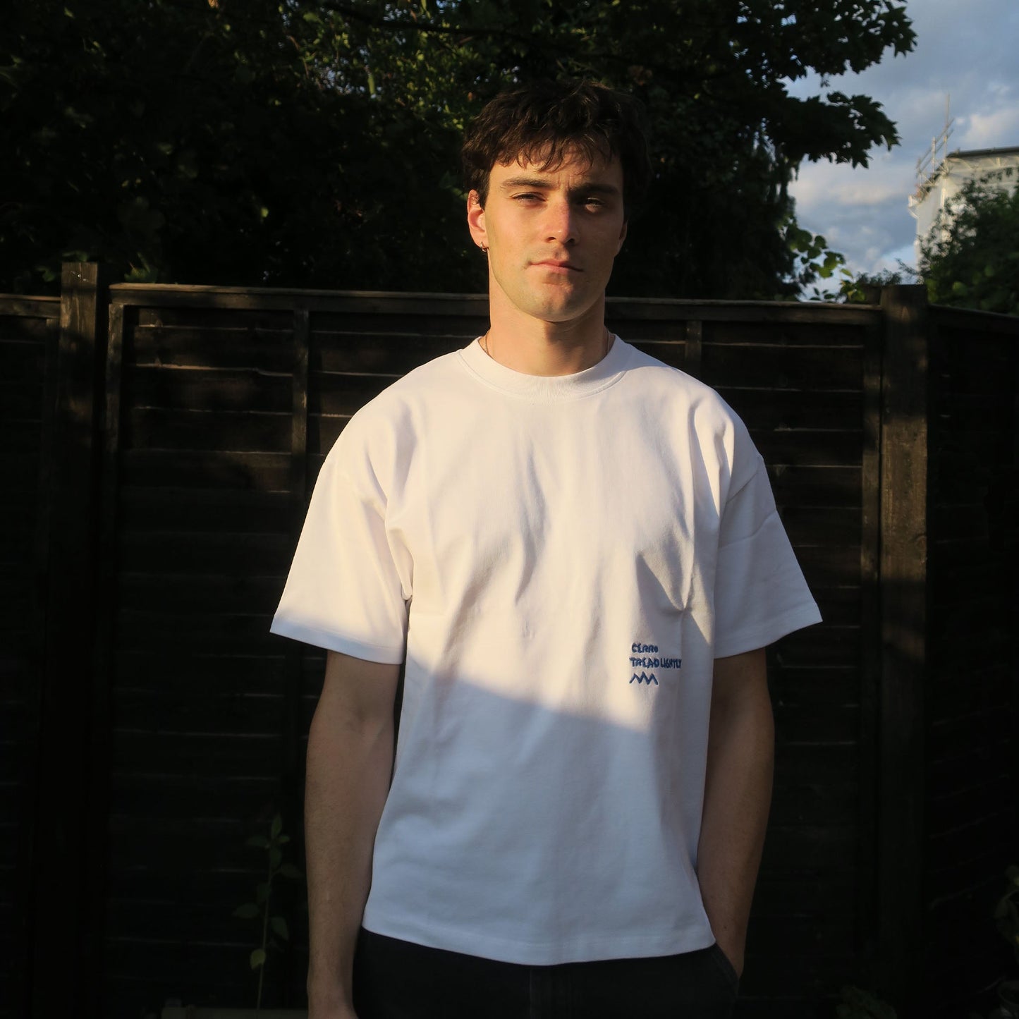 Person wearing a white t-shirt with embroidered blue text outdoors. 