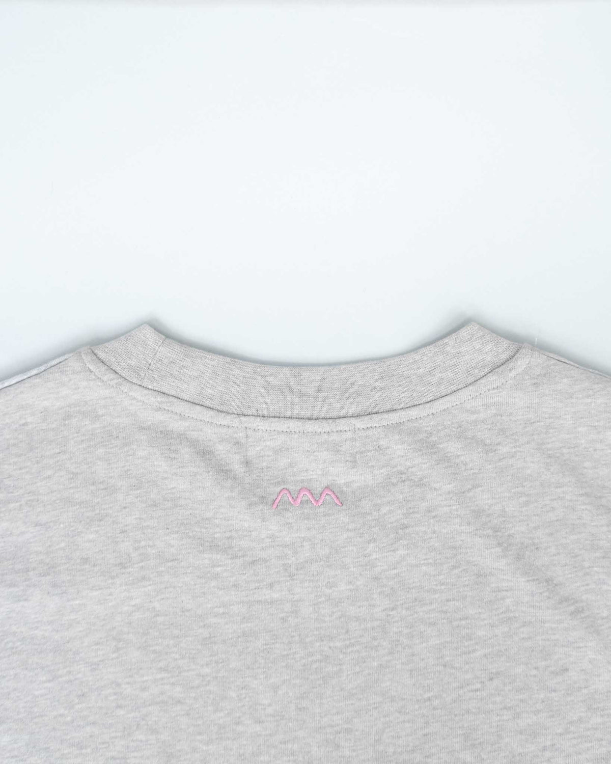 Close-up of the back of a light heather gray crewneck sweatshirt, featuring a small pink embroidered zigzag logo centered below the collar.