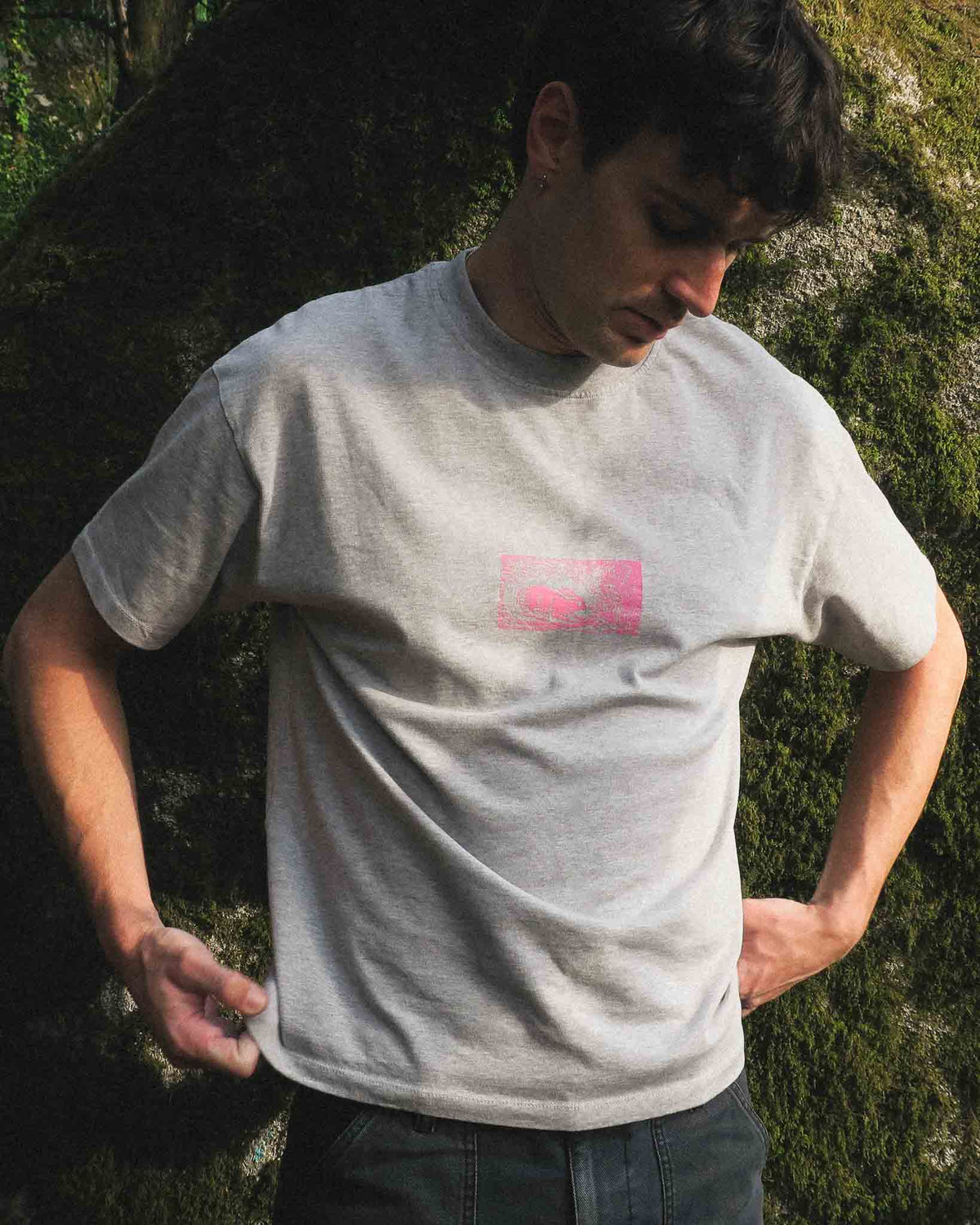 A man wearing a light gray crewneck t-shirt with a small pink graphic on the chest, standing outdoors against a mossy stone.