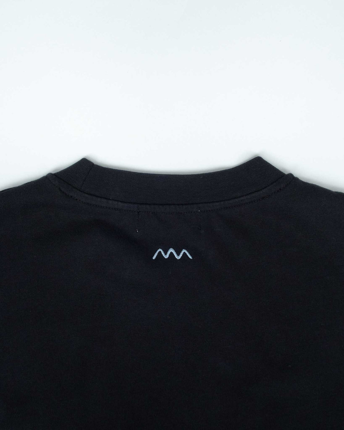 Close-up of the back collar of a black t-shirt featuring a small blue embroidered mountain-like logo on the center of the neck, set against a plain white background.