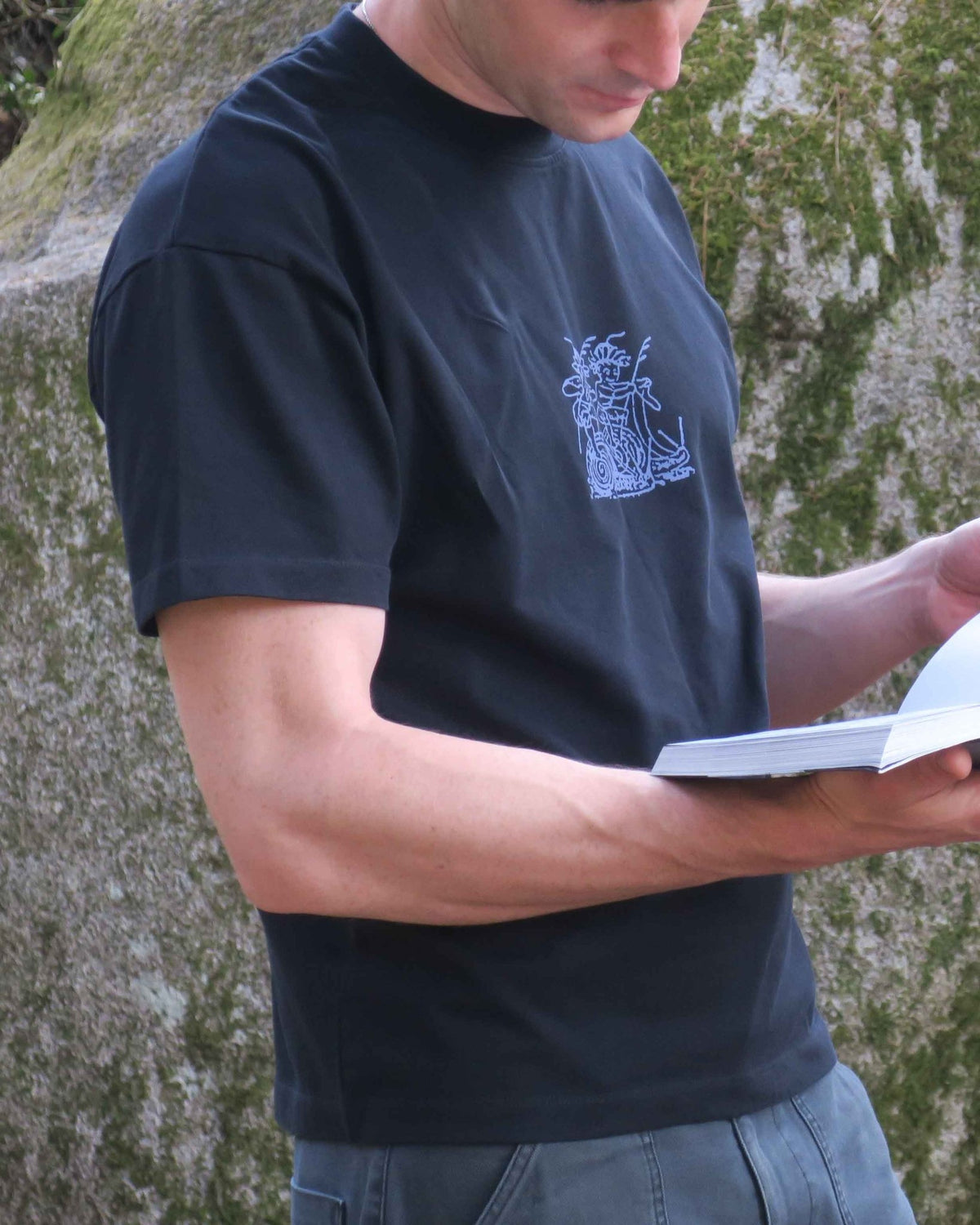 A man wearing a black crew-neck t-shirt with a small blue line-art graphic on the left chest, holding an open book in front of a mossy rock wall.