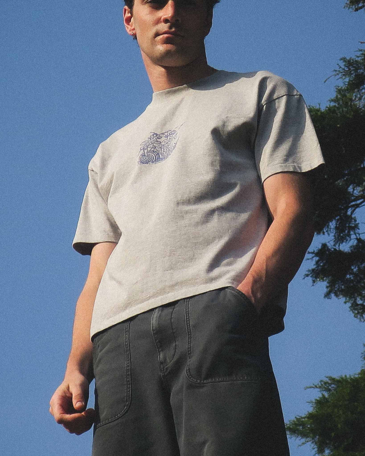 A man wearing a light beige crewneck t-shirt with a small blue graphic on the left chest, paired with dark gray cargo pants, photographed outdoors against a clear blue sky.