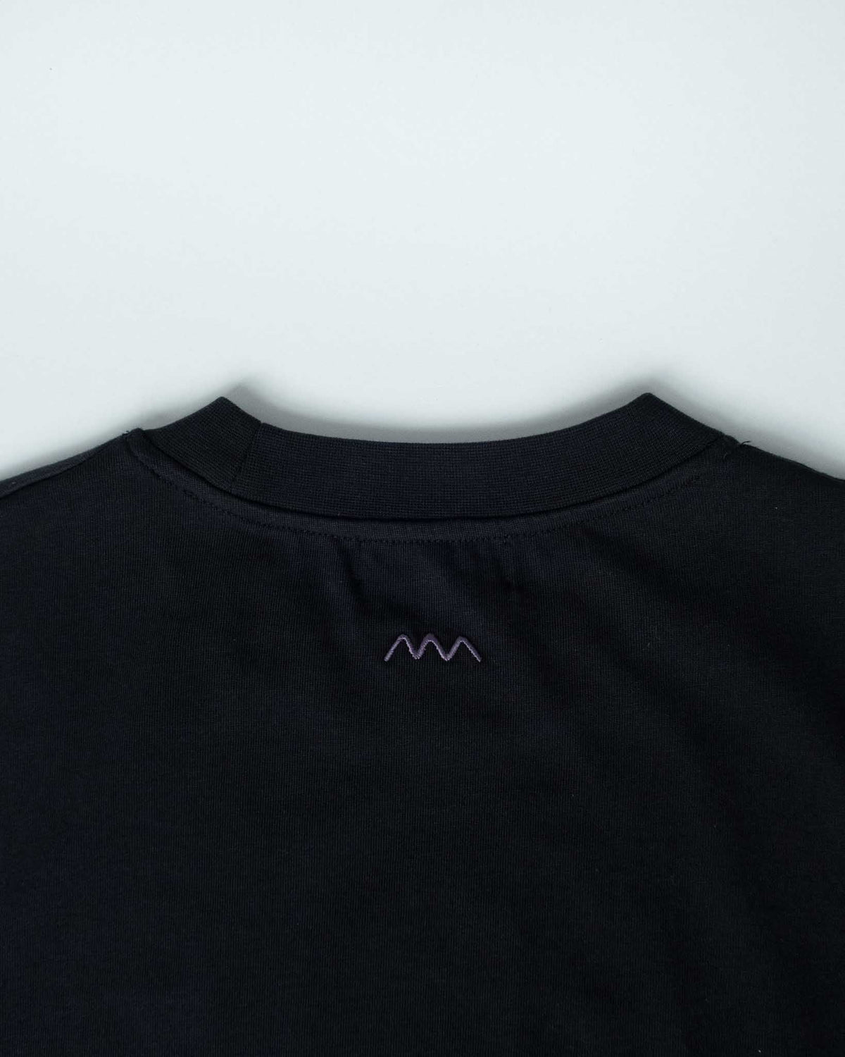 Close-up of the back collar of a black crewneck t-shirt, featuring a small purple embroidered mountain-like logo on the upper back.