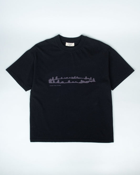 A black short-sleeve crewneck t-shirt with a dark gray horizontal graphic of a stone circle and the text 'COUNT THE STONES' printed across the chest, displayed flat against a light gray background.