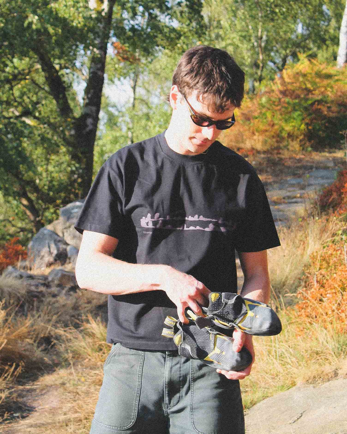 A man wearing a black t-shirt and sunglasses stands outdoors in a wooded area, holding a pair of black and gray climbing shoes.